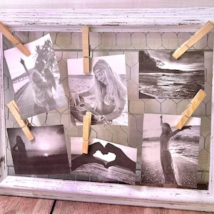 May include: A white wooden picture frame with a wire mesh backing holds a collection of black and white photographs. The photos are secured with wooden clothespins. The photos depict various scenes, including a woman playing a guitar, a woman standing in a field, and a woman walking on a beach.