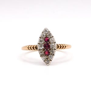 May include: A gold ring featuring a marquise-shaped setting with three central red rubies arranged vertically, surrounded by a halo of small clear white stones. The band has a textured, ribbed design on both sides of the central gemstone cluster.