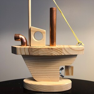 May include: A wooden toy boat with a copper pipe, a copper mast, and a yellow rubber band. The boat has a small wooden cabin with a round hole in the front. The boat is sitting on a black surface.