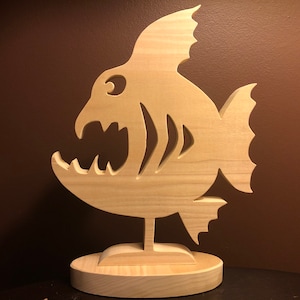 May include: A wooden cutout of a fish with a large open mouth and sharp teeth. The fish is standing on a wooden base.