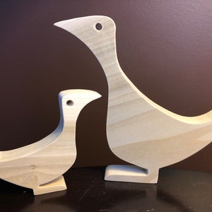 以下が含まれることがあります： 2つの木製の鳥の彫刻。1つは他方より大きく、明るい木で作られており、シンプルで抽象的なデザインです。鳥は前を向いて、頭を上向きに傾けています。