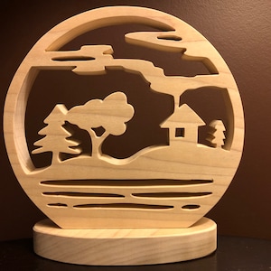 Puede incluir: Un recorte de madera de una escena de paisaje con una casa, árboles y nubes. El recorte está montado sobre una base de madera.