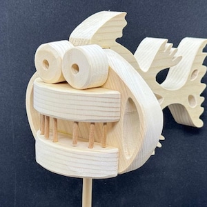 May include: A wooden toy fish with a toothy grin, large eyes, and a fin on the back. The fish is unpainted and made of light wood.