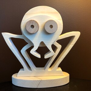 Könnte beinhalten: Eine Holzspinne-Figur mit hellbrauner Oberfläche. Die Spinne hat einen großen Kopf mit zwei runden Augen und acht Beinen. Die Spinne steht auf einem kleinen Holzsockel.