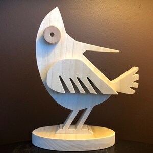 Könnte beinhalten: Eine Holzvogelskulptur mit einem skurrilen Design. Der Vogel ist in einer natürlichen Holzfarbe bemalt und hat ein großes, rundes Auge. Der Vogel steht auf einem Holzsockel.