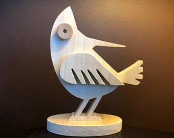 May include: A wooden bird sculpture with a whimsical design. The bird is painted in a natural wood color and has a large, round eye. The bird is standing on a wooden base.