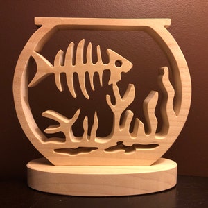 May include: A wooden fishbowl sculpture with a fish skeleton inside and seaweed. The fishbowl is on a wooden base.