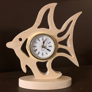 May include: A wooden clock shaped like a fish with a white face and black Roman numeral markings. The clock has a gold-colored frame and hands.