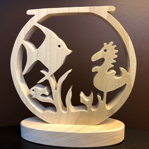 May include: A wooden cutout of a fish tank with a seahorse, two fish, and seaweed. The tank is made of light wood and is standing on a wooden base.