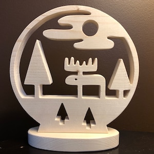 May include: A wooden, circular decorative piece featuring a carved forest scene. The design includes a moose, trees, a sun, and clouds. The piece is mounted on a round base and is made of light-colored wood.