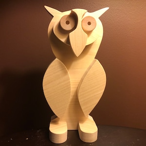 Puede incluir: Una escultura de búho de madera con ojos grandes y un pico. El búho está pintado en un color madera claro.