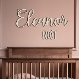 May include: A wooden crib with a mattress and a pink pillow is in a room with a pink wall. Above the crib, the name "Eleanor Rose" is displayed in a cursive font. A woven basket with a stuffed animal is next to the crib.