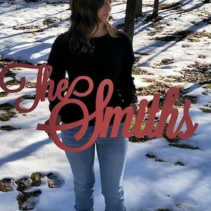 May include: A red wooden sign with the text "The Smiths" in a cursive font. The sign is being held by a person in front of a snowy background.