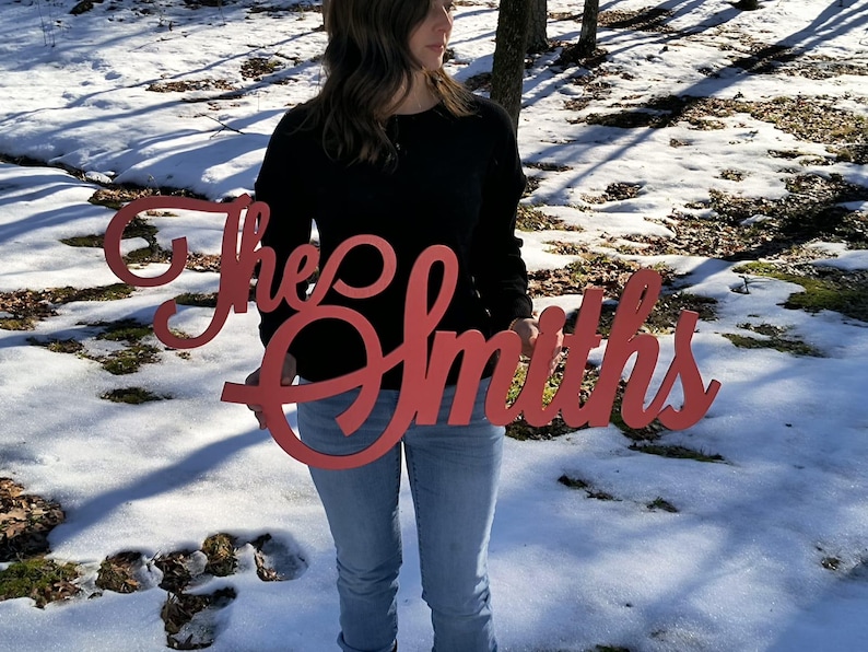 May include: A red wooden sign with the text "The Smiths" in a cursive font. The sign is being held by a person in front of a snowy background.