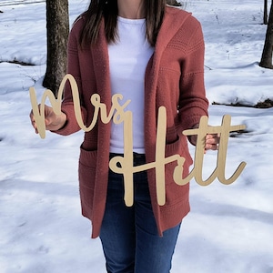 May include: A person holding two wooden signs that say "Mrs. Hitt" in a cursive font. The signs are light brown and have a natural wood finish.