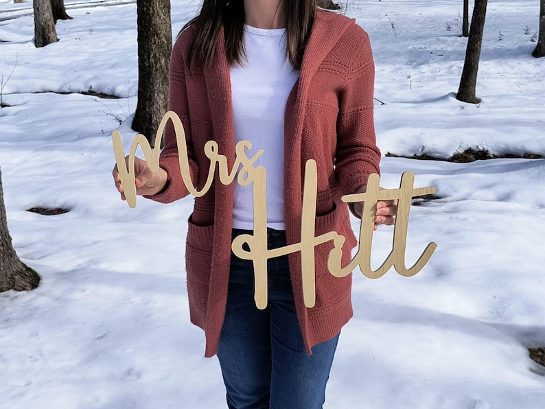 May include: A person holding two wooden signs that say "Mrs. Hitt" in a cursive font. The signs are light brown and have a natural wood finish.