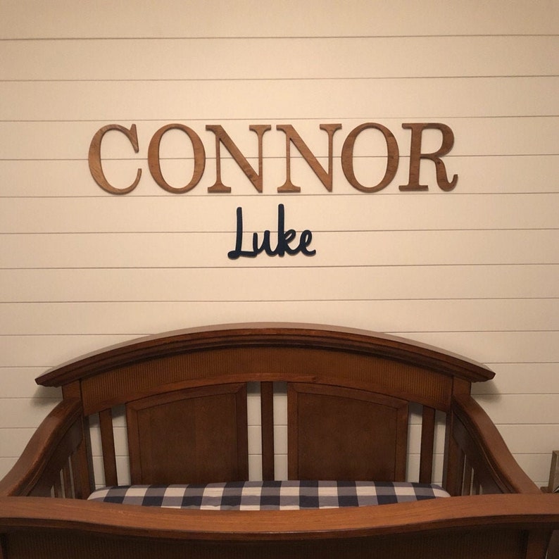 May include: Wooden letters spelling the name "CONNOR" in brown, with the name "Luke" in blue cursive lettering below, above a wooden cot with a black and white checked mattress.