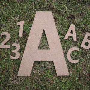 May include: A collection of large, unfinished cardboard letters and numbers arranged on a bed of green grass. The set includes the letters A, B, and C, along with the numbers 1, 2, and 3. The neutral color of the cardboard contrasts with the green background.