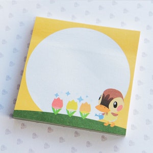 May include: Square yellow sticky note pad with a large white circle for writing. Features a cartoon duck watering flowers. The background is yellow, with a green strip at the bottom. The pad is on a white surface.