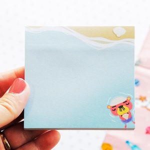 May include: A square sticky note with a light blue background, featuring a cartoon otter floating in water. The top edge has a beach scene with a wave and a cloud. The otter wears a white hat and purple shorts.