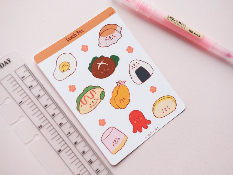 以下が含まれることがあります： かわいい顔をしたさまざまな日本の食べ物を描いたステッカーシート。ステッカーは白地に桃色の縁取りが施されています。シートの上部には「Lunch Box」の文字があります。
