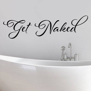 May include: A black wall decal featuring the cursive script text Get Naked positioned above a white freestanding bathtub with a chrome faucet fixture. The lettering is applied directly to the wall surface in a decorative, flowing font style.
