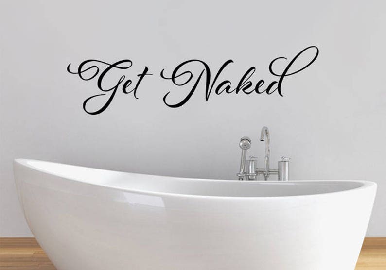 May include: A black wall decal featuring the cursive script text Get Naked positioned above a white freestanding bathtub with a chrome faucet fixture. The lettering is applied directly to the wall surface in a decorative, flowing font style.
