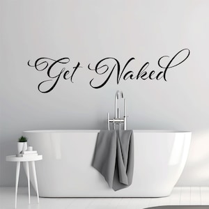 May include: A black vinyl wall decal featuring the text Get Naked in a cursive script font. The lettering is applied to a plain wall above a white bathtub with a chrome faucet and a gray towel draped over the side, next to a small white table.