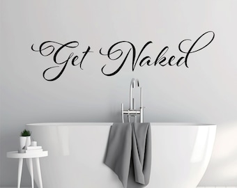May include: A black vinyl wall decal featuring the text Get Naked in a cursive script font. The lettering is applied to a plain wall above a white bathtub with a chrome faucet and a gray towel draped over the side, next to a small white table.