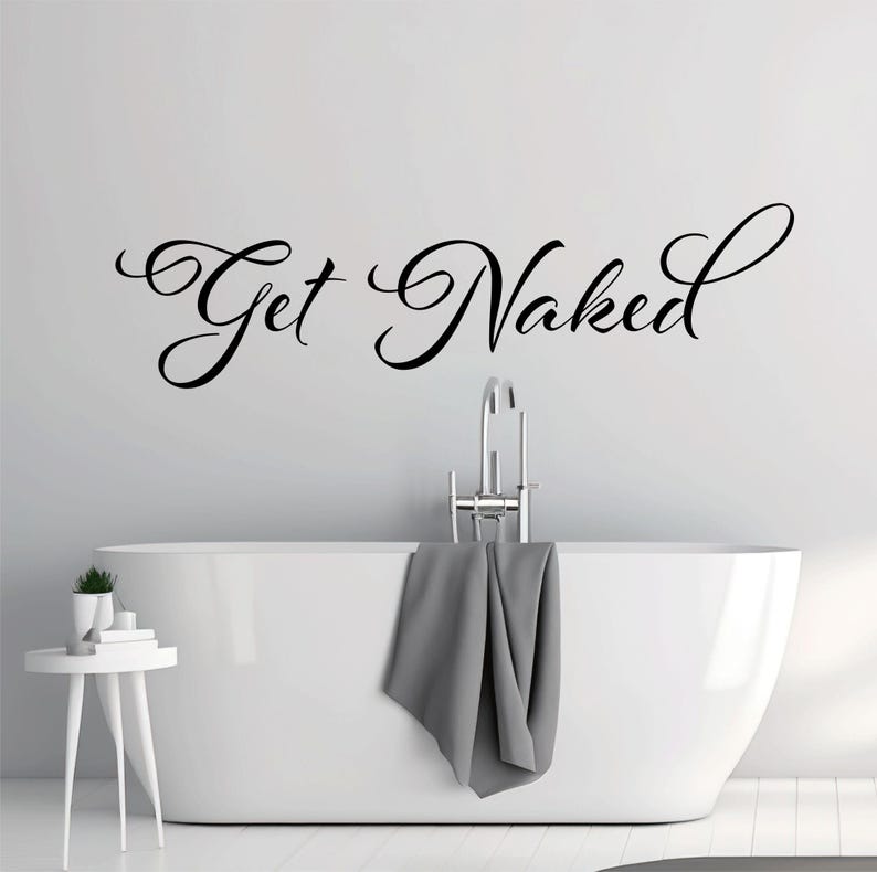 May include: A black vinyl wall decal featuring the text Get Naked in a cursive script font. The lettering is applied to a plain wall above a white bathtub with a chrome faucet and a gray towel draped over the side, next to a small white table.