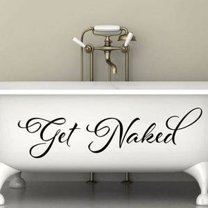 May include: A white clawfoot bathtub featuring the black cursive text Get Naked centered on the front side. The tub has four rounded feet and is positioned in front of a metallic faucet fixture with white handles and a central spout.