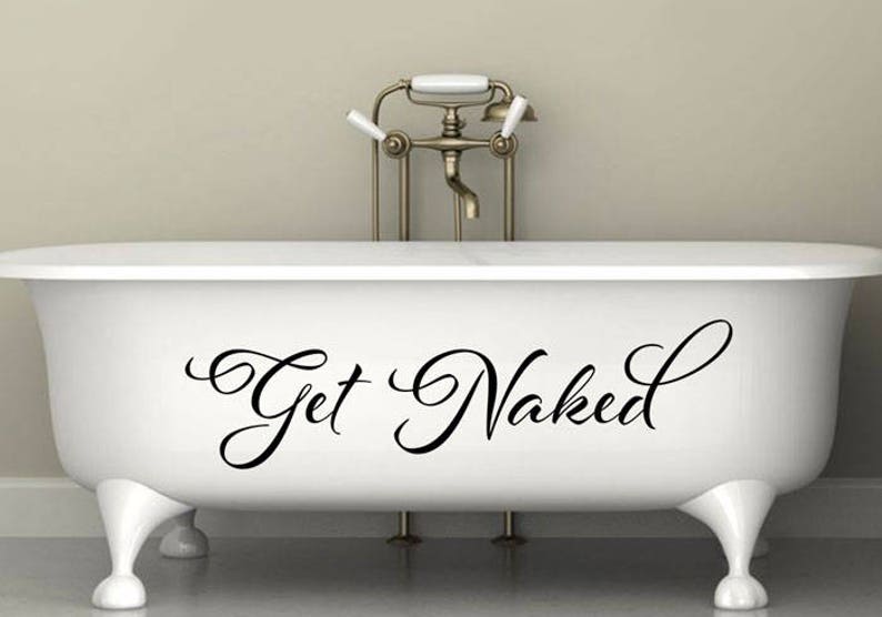 May include: A white clawfoot bathtub featuring the black cursive text Get Naked centered on the front side. The tub has four rounded feet and is positioned in front of a metallic faucet fixture with white handles and a central spout.