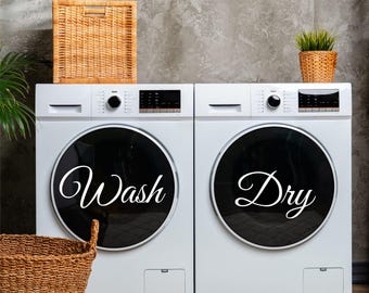 May include: Two white washing machines side-by-side with black round doors. The left machine has the word "Wash" in white cursive on the door. The right machine has the word "Dry" in white cursive on the door.