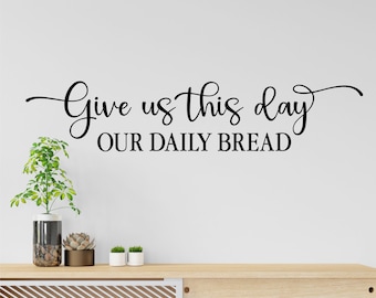 May include: A black and white wall decal with the text "Give us this day our daily bread". The decal is on a white wall above a wooden countertop with plants and a vase.