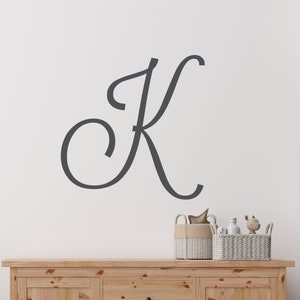 May include: A grey cursive letter K wall decal on a white wall above a wooden dresser with two baskets.