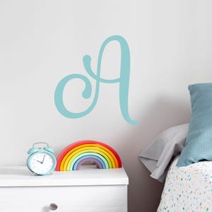 May include: A light blue cursive letter A wall decal above a rainbow-colored wooden stacking toy on a white dresser.