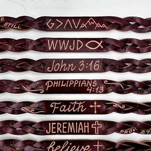 May include: A set of eight leather bracelets with different engraved messages, including "Be Still", ">AVA", "WWJD", "John 3:16", "Philippians 4:13", "Faith", "Jeremiah", and "Believe". The bracelets are braided and have a dark brown color.