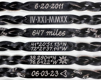 May include: Black leather bracelet with multiple engraved lines of text, including dates, coordinates, and the words "Under the Same Sky".