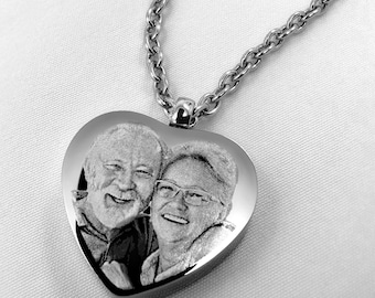 Op de afbeelding: Een zilverkleurige hartvormige hanger met een lasergegraveerd zwart-wit fotoportret van een glimlachend ouder echtpaar. De hanger is bevestigd aan een bijpassende zilverkleurige metalen schakelketting met een gepolijste afwerking.