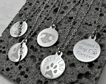 May include: Five silver necklaces with round pendants featuring paw prints and personalized engravings. The pendants read "Cali", "Jalapeño", "Mama's Boy Forever", and one with a heart and paw print.