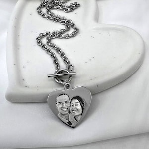 May include: A silver heart-shaped pendant necklace with a photo of a couple engraved on the front. The necklace has a toggle clasp and a silver chain.
