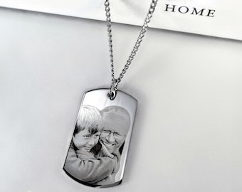 Op de afbeelding: Een zilveren ketting met een zilveren hondenpenning hanger. De hondenpenning is gegraveerd met een zwart-witte foto van twee mensen.