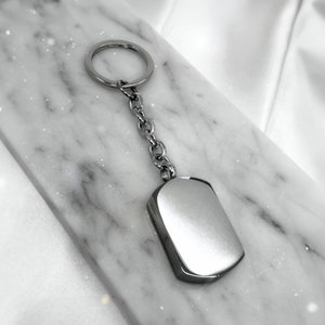 May include: A silver keychain with a round ring and a rectangular tag. The tag is smooth and has a shiny finish.