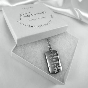 May include: A silver dog tag keyring with the inscription "Until we meet again at the rainbow bridge" and paw prints. The keyring is in a white box with the words "Karved Jewellery Studio" printed on the lid.