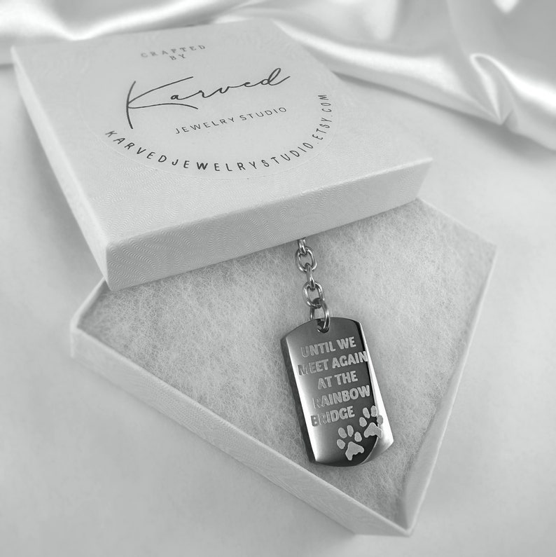 May include: A silver dog tag keyring with the inscription "Until we meet again at the rainbow bridge" and paw prints. The keyring is in a white box with the words "Karved Jewellery Studio" printed on the lid.