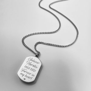 May include: A silver-colored dog tag pendant on a chain. The tag is rectangular with rounded corners and features engraved text: "Salazar Morales 1955-2021 Forever in my heart" with a small heart symbol. The chain is a curb style.