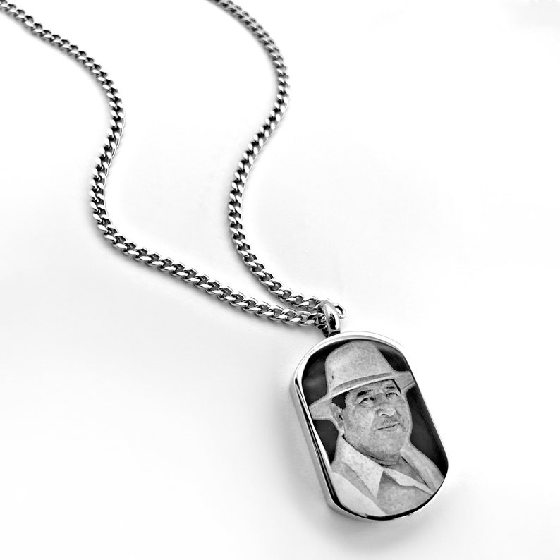 May include: Silver-toned dog tag pendant necklace featuring a black and white portrait of a person wearing a hat. The pendant hangs from a curb chain. The image is set against a white background.