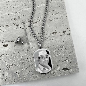 May include: A silver-colored memorial pendant necklace with a rectangular tag featuring a black and white portrait of a man wearing a hat. The necklace includes a chain, a small funnel, and a filling tool.