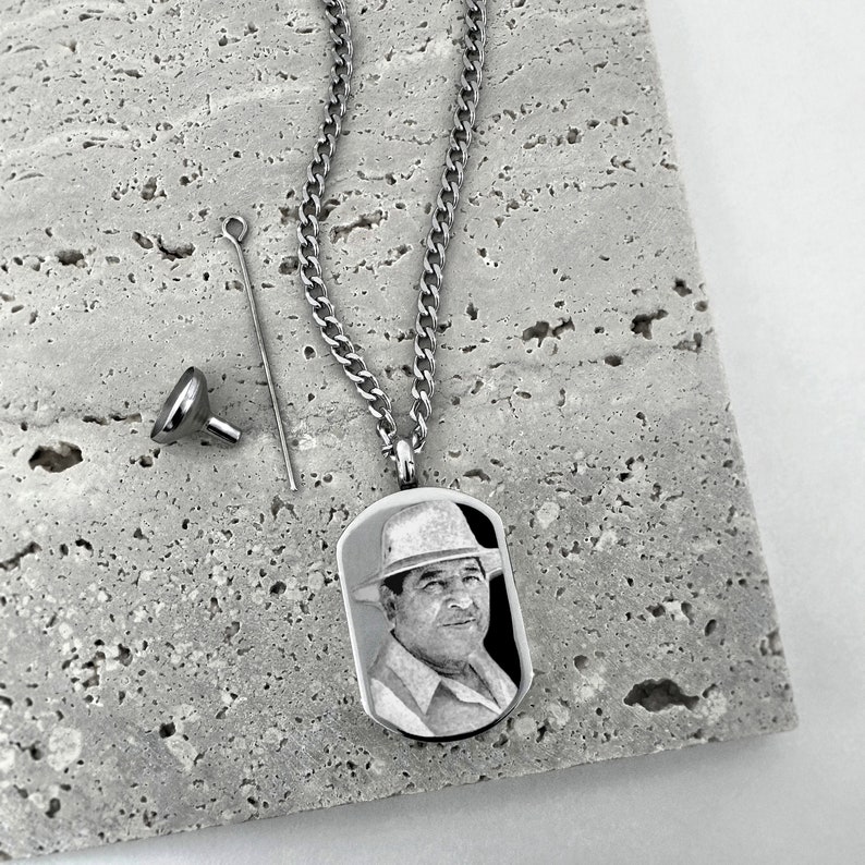 May include: A silver-colored memorial pendant necklace with a rectangular tag featuring a black and white portrait of a man wearing a hat. The necklace includes a chain, a small funnel, and a filling tool.