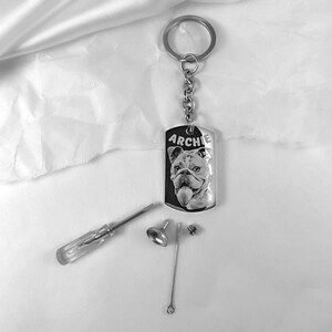 May include: Silver keyring with a black and white photo of a dog engraved on a rectangular tag. The tag says "ARCHIE". The keyring comes with a small screwdriver, a metal ring, and a small metal cap.
